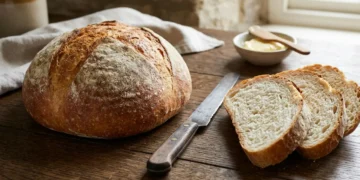 Tradycyjny bochenek chleba pszennego i pokrojone kromki na drewnianym stole, ilustrujące kaloryczność białego pieczywa.